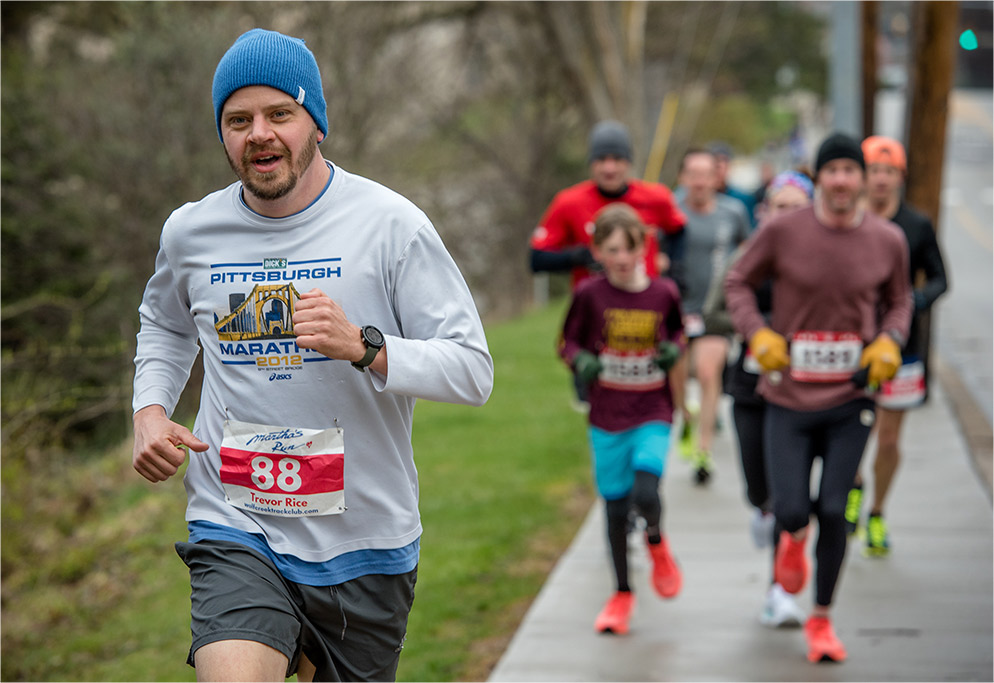 A man runs on the sidewalk with a "Martha's Run" bib and Pittsburgh Marathon 2012 top and beanie, with a trail of runners behind him in a blur