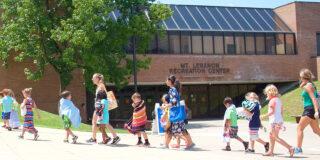 Parents and children walk in a single file line, wrapped in beach towels, in front of the Mt. Lebanon Recreation Center on a sunny day. They head toward the Mt. Lebanon pool.