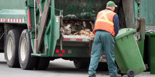 A man with a bright orange vest pushes a green garbage can toward the back of a garbage truck.