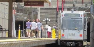 The T pulling into the Mt. Lebanon station with a group of people walking towards it to get on. The sign at the stop reads 