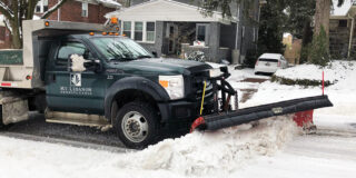 A Mt. Lebanon public works truck with a plow on the front drives down a street, plowing snow.