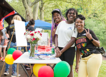 Three black females smile for the camera, one is giving the peace sign, standing by a table outside surrounded by balloons
