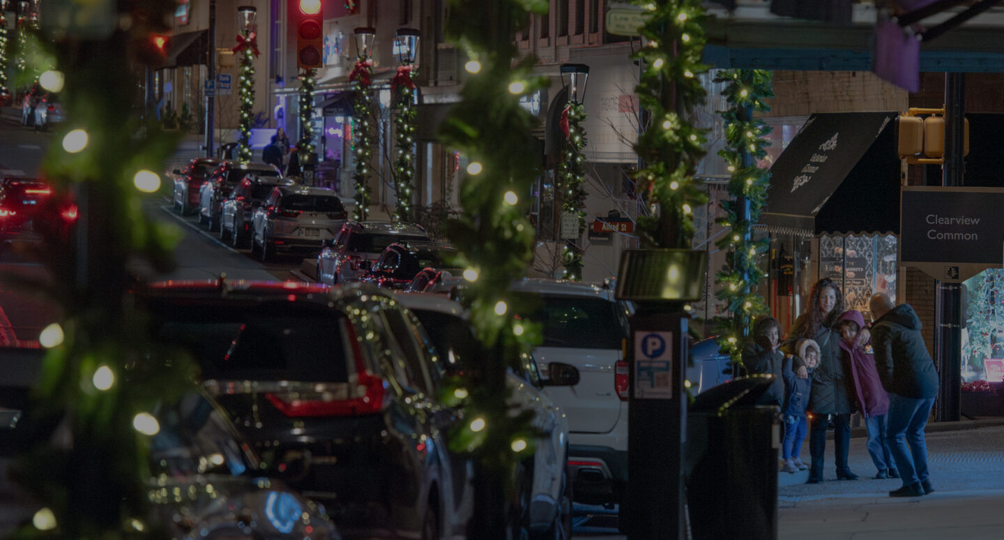 Washington Road lined with parked cars at night with lamp posts wrapped in green garland with white lights and red bows at the top and a family posing for a photo on the sidewalk in the bottom right.