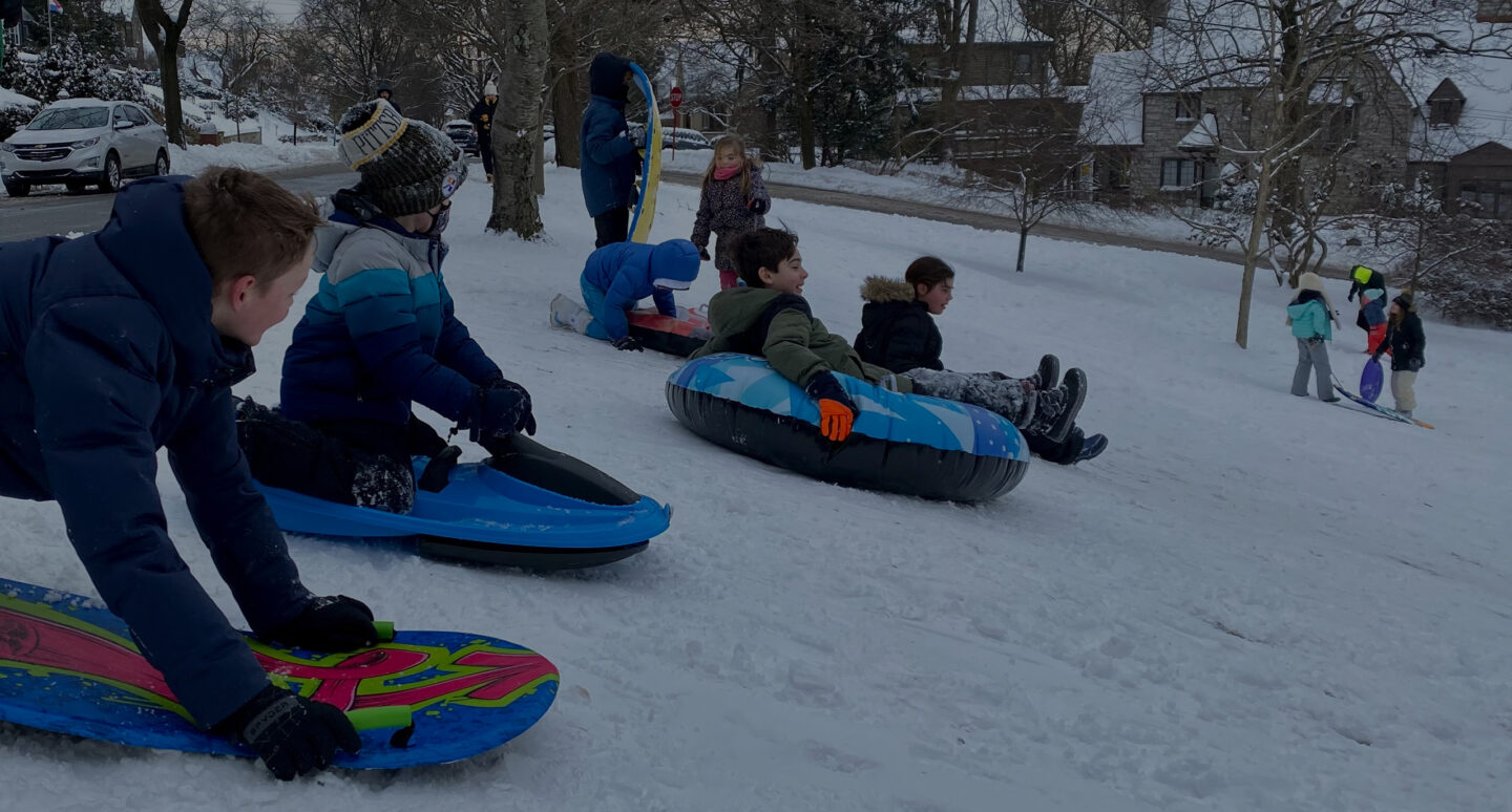 A group of kids on sleds at the top of a snow covered hill with homes and trees in the background.