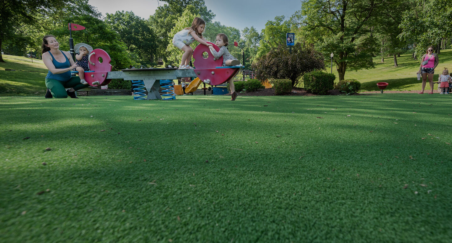 A playground scene with children playing on a seesaw. A woman assists a toddler on one side, while two kids balance on the other.