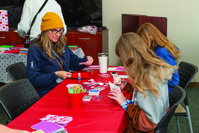 Image of a woman and two children making valentines at a table with a red tablecloth, during a service project