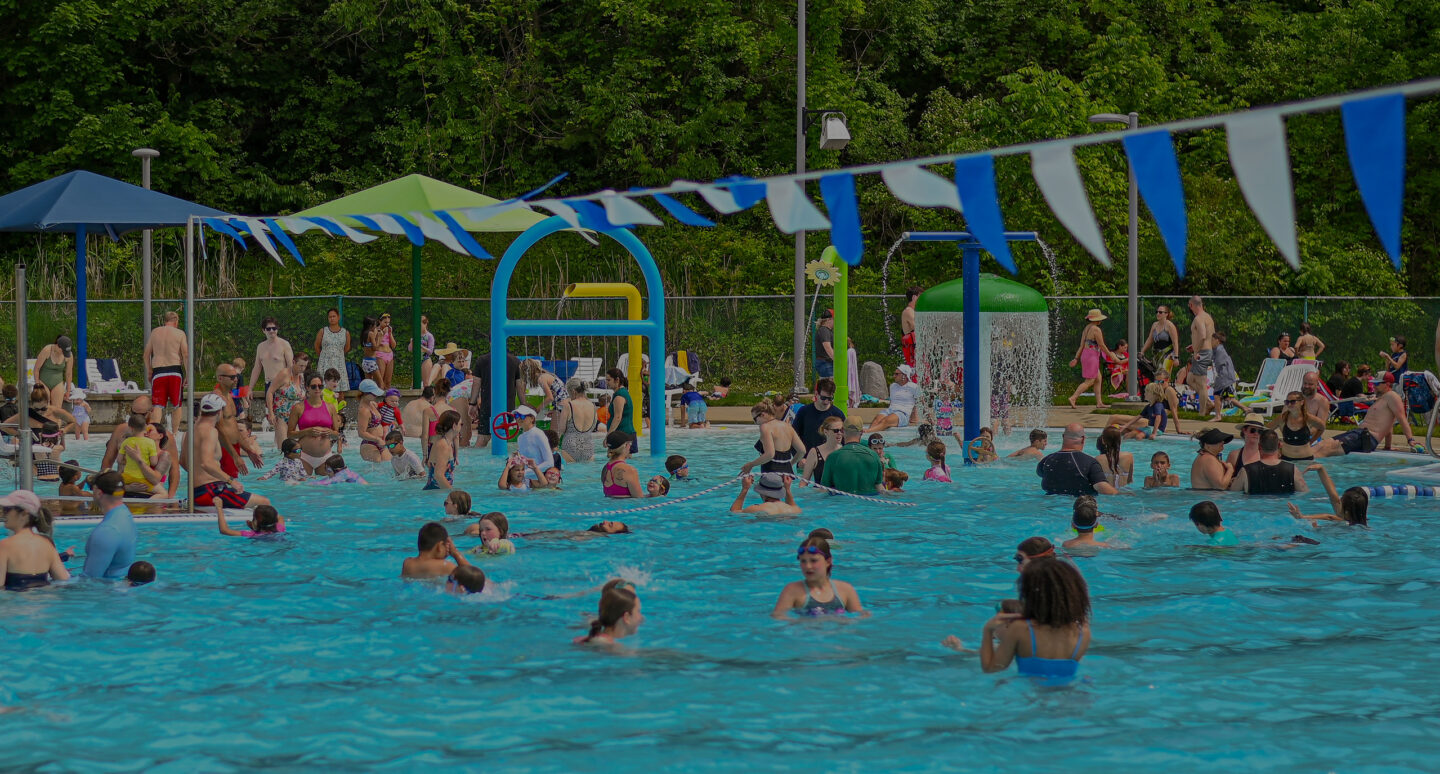 A crowd of people in a large swimming pool enjoying their day. Umbrellas and water features can be seen in the background.